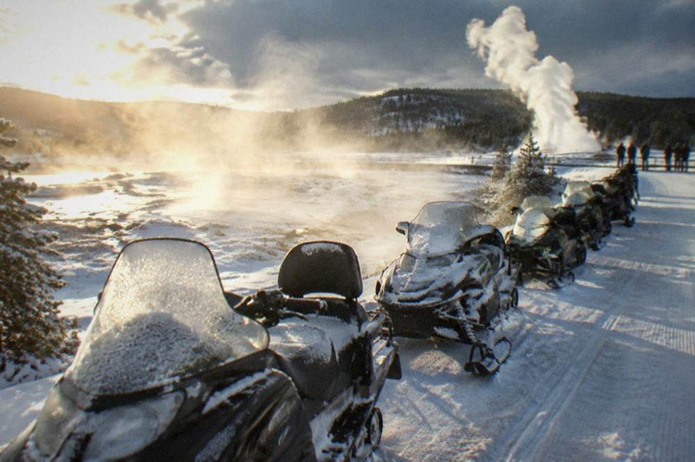 Row of parked snowmobiles lined up on a groomed snow road beside steam rising from geothermal ground