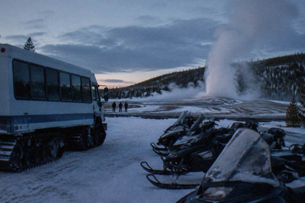 A snowcoach and parked snowmobiles on a groomed white road beside steaming thermal pools