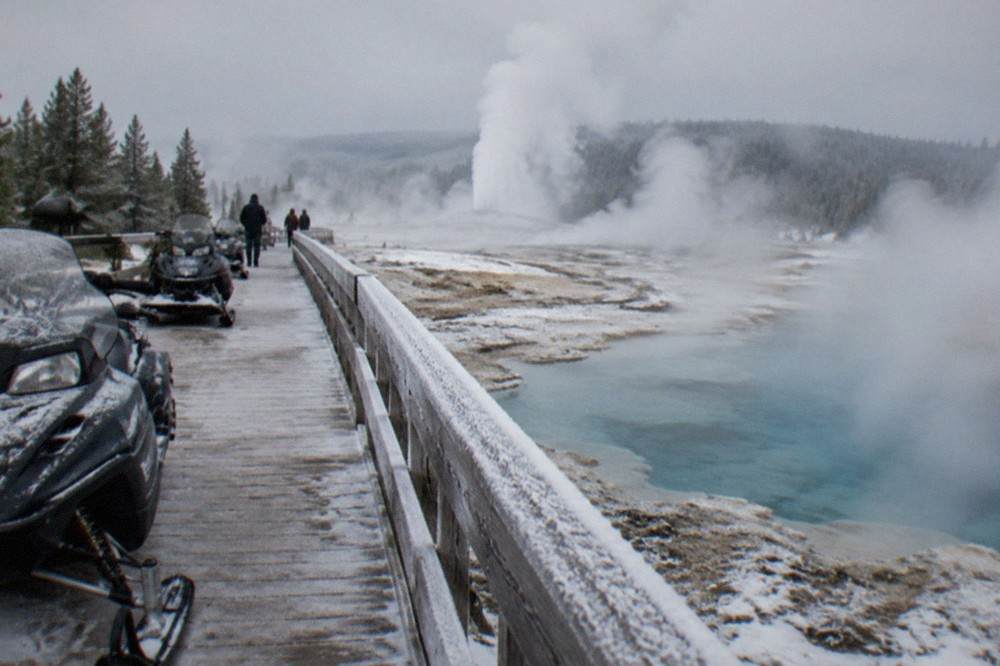 Snowmobiles parked in a line beside a snowy boardwalk with steam rising from geothermal pools.