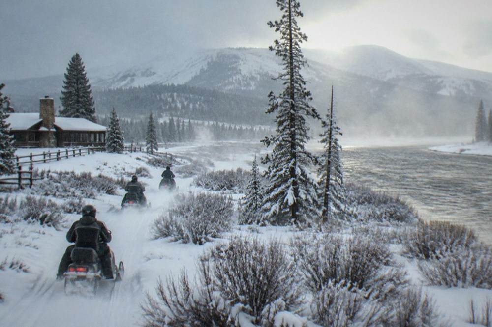 A guided snowmobile group riding single file along a groomed snow road between frosted pine trees.