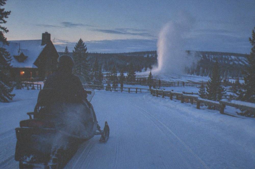 A snowmobile rider stops on a groomed road with Old Faithful erupting in the distance