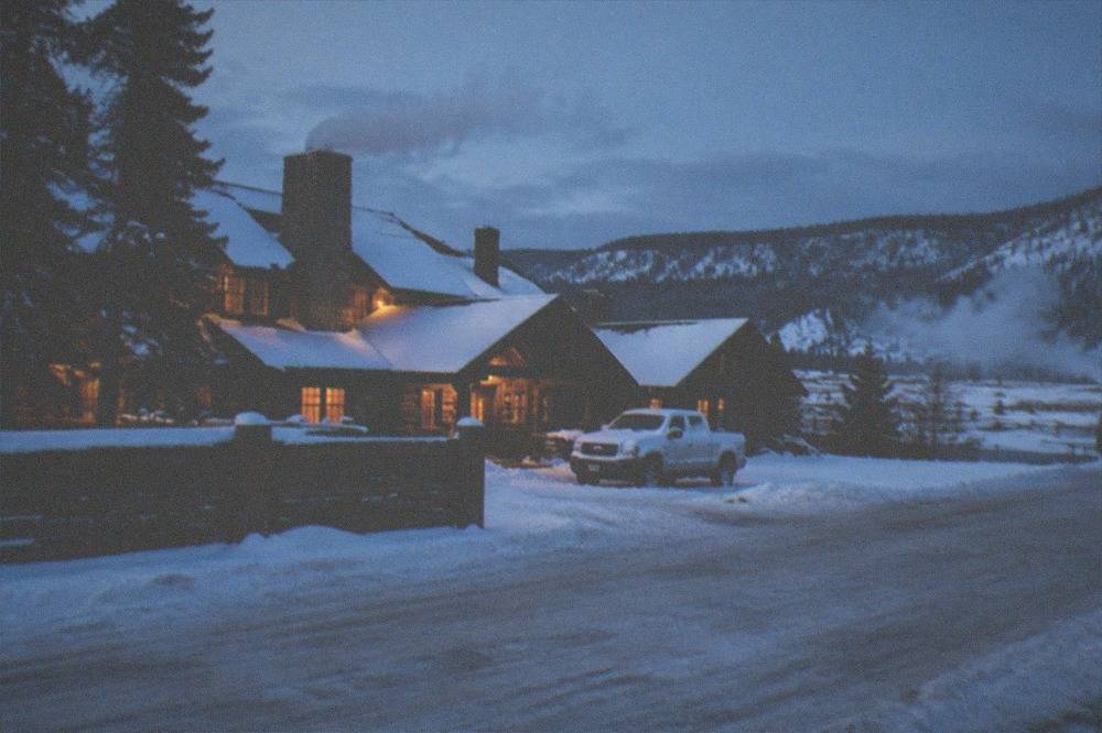 Warm lodge lights glowing at dusk against a snowy town street and dark pines