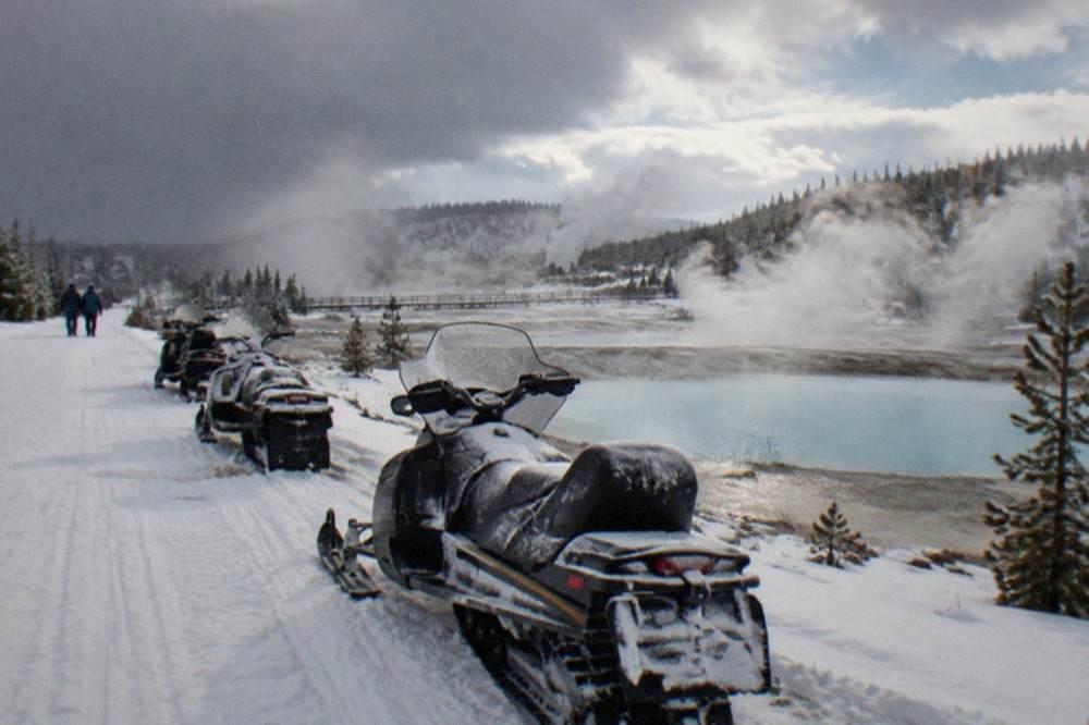 Line of parked snowmobiles on a groomed white road beside steaming thermal pools