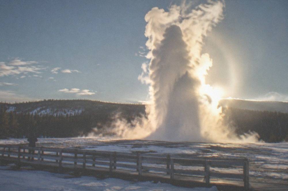 Geyser erupting in a tall plume above snow-covered ground and empty boardwalks