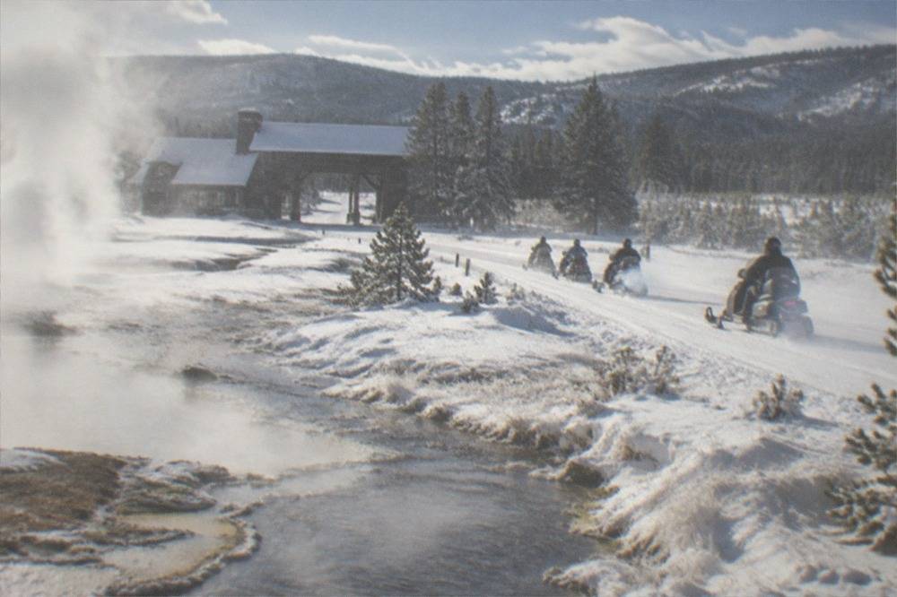 Line of snowmobiles traveling a groomed white road past steaming geothermal pools in a snowy valley