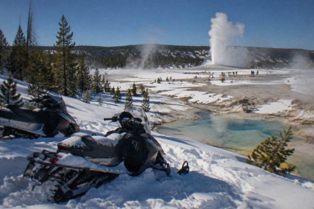 Snowmobiles parked in deep snow beside steaming geothermal pools under a cold blue sky