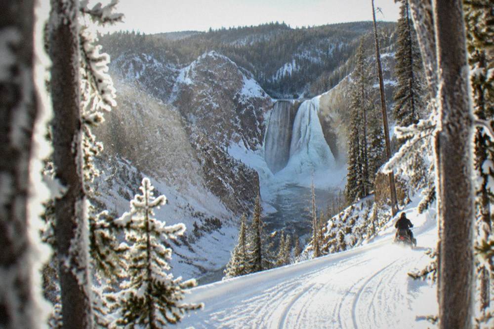 A snowmobile track winding along a groomed white road toward a frozen canyon with an icy waterfall