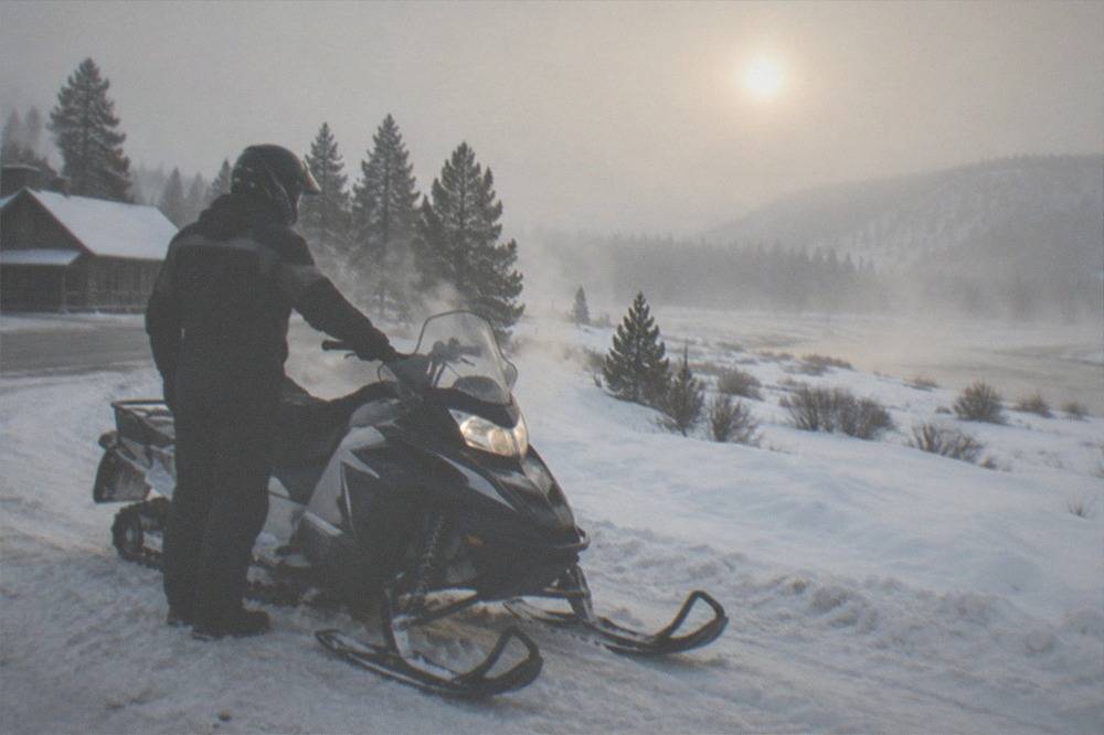 A snowmobile rider in an insulated suit and full-face helmet pausing on a wide snowy road beside frozen trees
