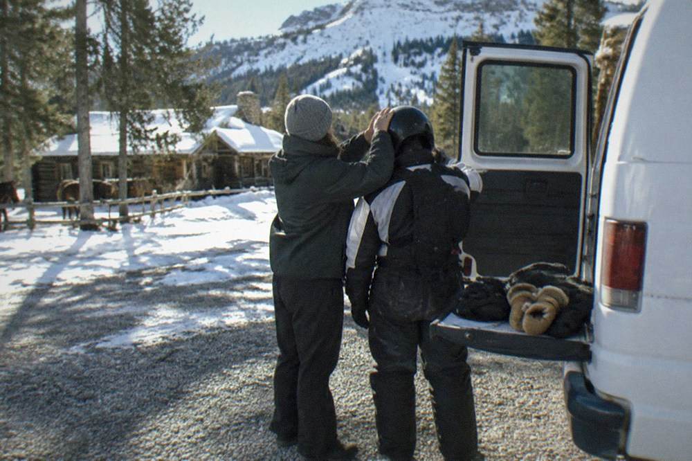 A snowmobile guide fitting a helmet for a rider beside a van in a snowy parking area
