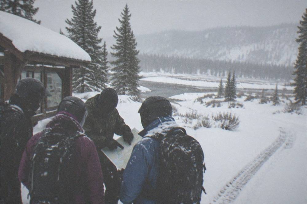 Riders in insulated snowmobile suits gathered around a guide during a safety briefing in falling snow