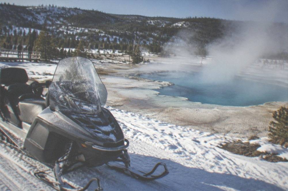 Snowmobile parked beside steaming thermal pools under a cold blue winter sky in Yellowstone