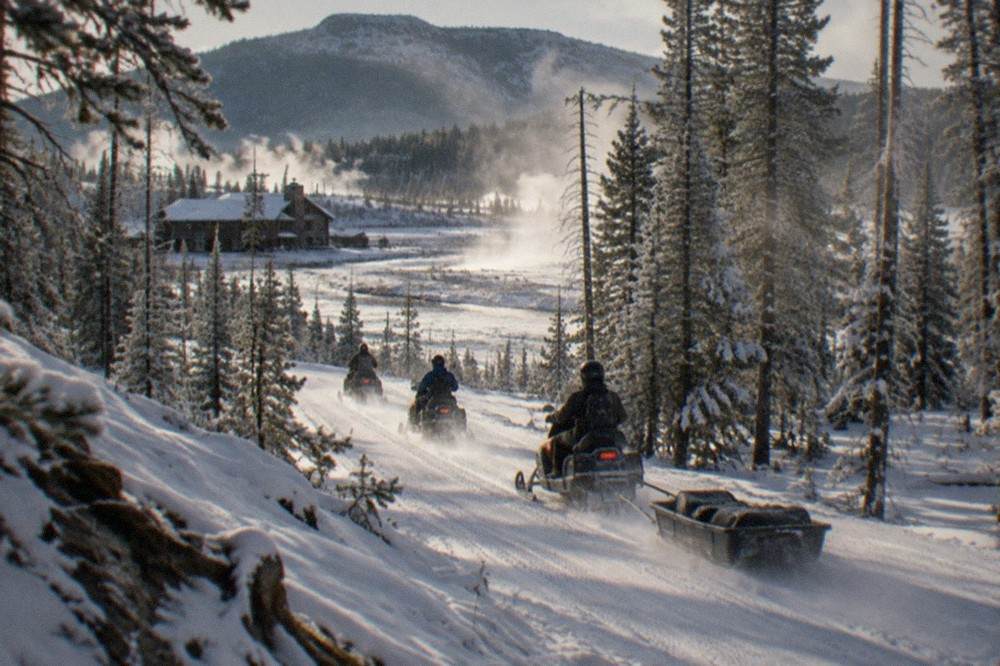 A guided line of snowmobiles traveling along a groomed snow-covered road through frosted pine forest