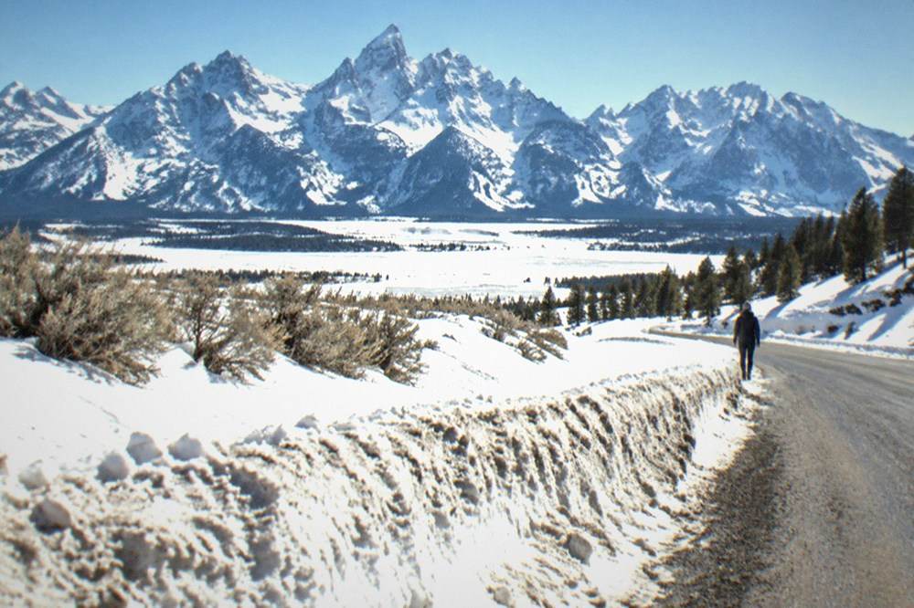 A plowed winter highway running along a snow-covered valley with jagged mountain peaks rising behind