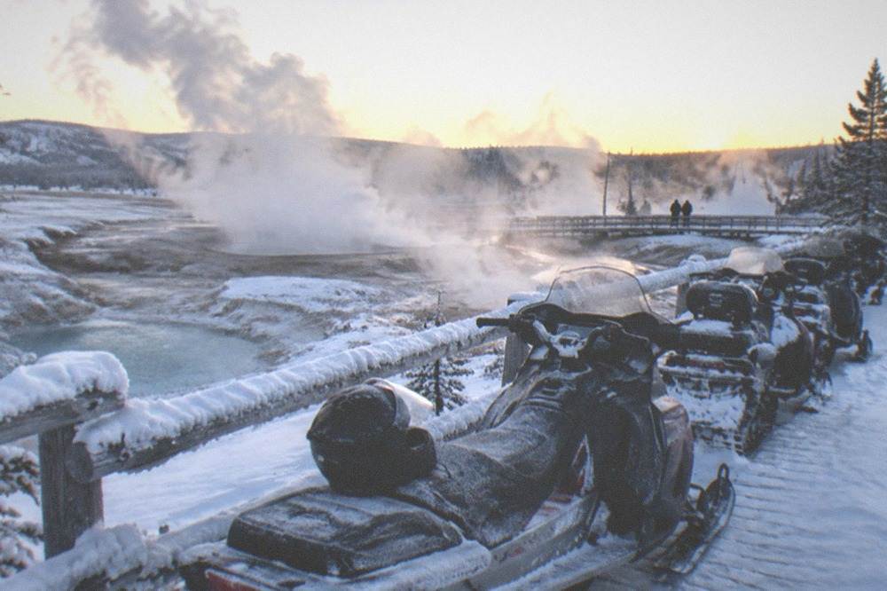 Snowmobiles parked in a line beside a snow-covered boardwalk with steam rising in the distance