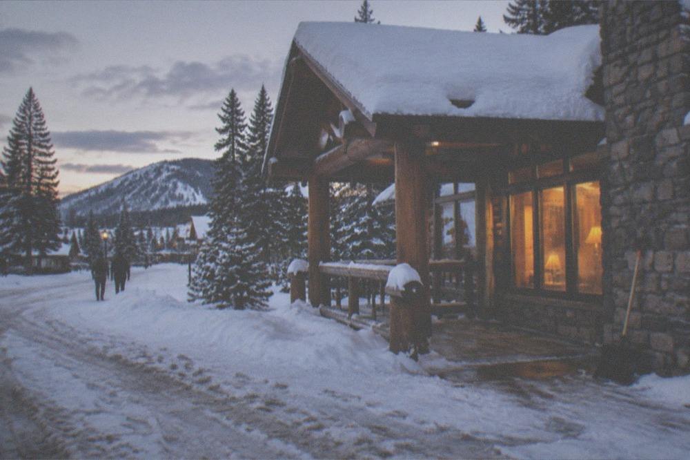 A snow-covered wooden lodge porch at dusk with warm light in the windows