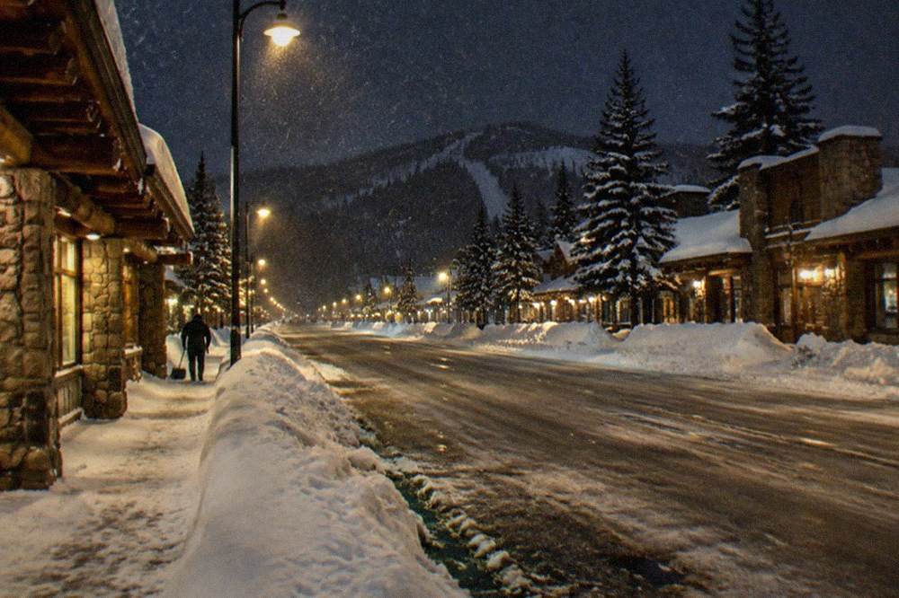 Small winter town street at dusk with warm shop lights and deep snow banks along the sidewalks