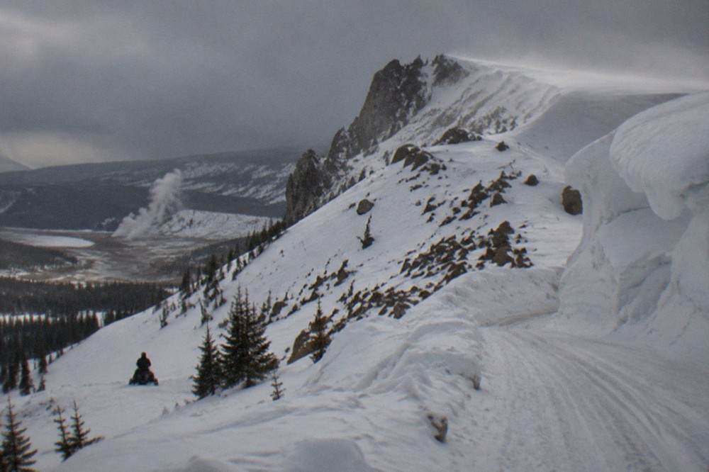 Steep snow-packed mountain road above timberline with cornices of wind-blown snow on the ridgeline.
