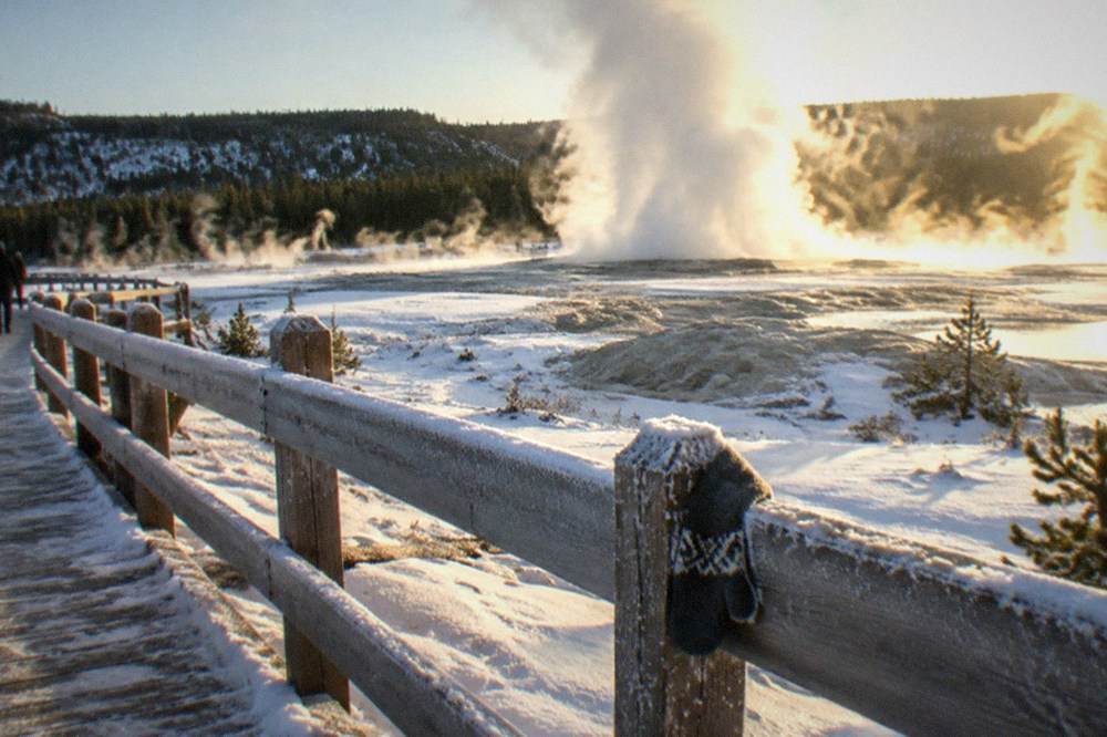 Geothermal steam rising from a snow-covered geyser basin beside a boardwalk in winter.