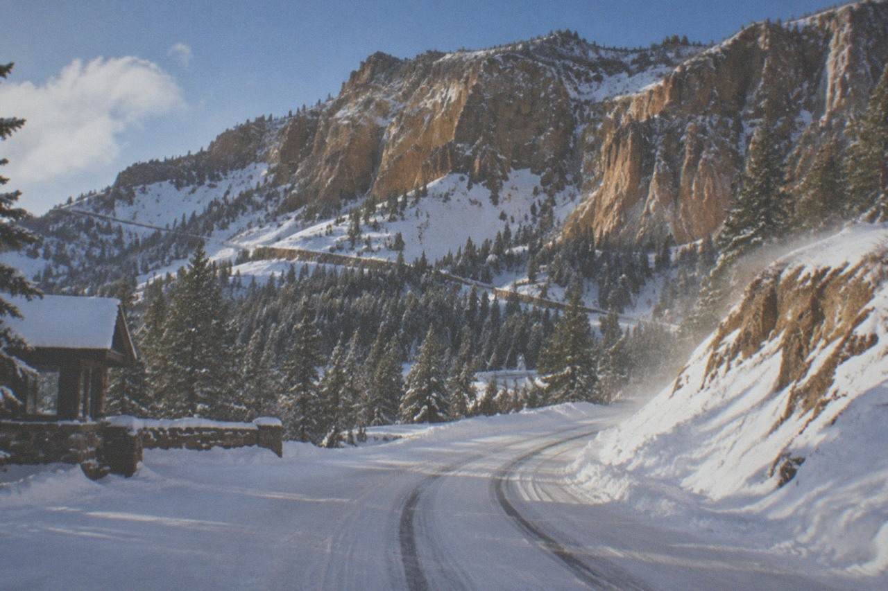 Snow-covered mountain pass road winding through pine forest under a cold blue sky.
