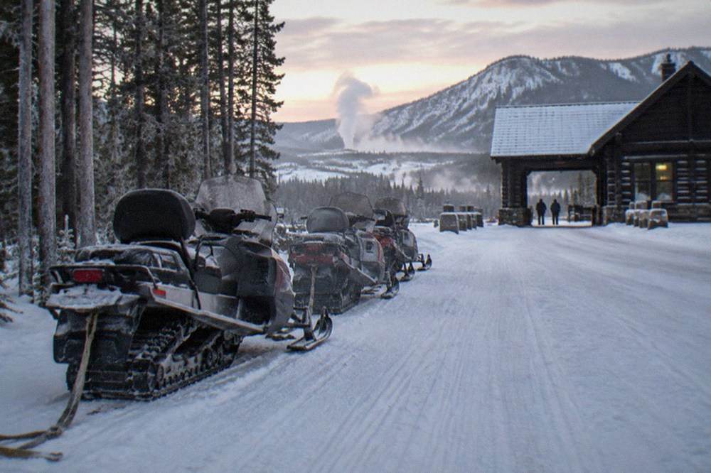 A line of parked snowmobiles on a groomed snow road beside frosted pine trees at dawn
