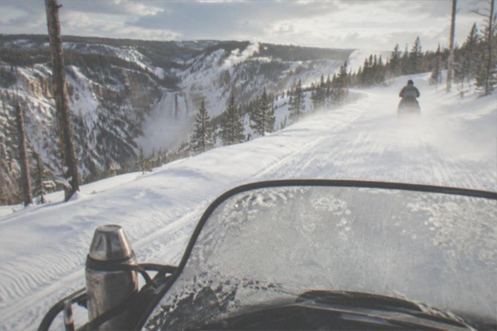 A snowmobile dashboard and handlebars seen from a rider's position on a wide groomed snow road