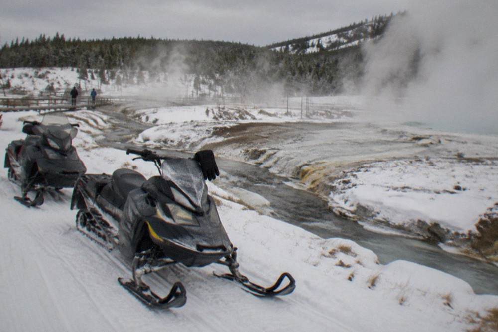 Two snowmobiles parked on a groomed snow road beside steaming hot springs in a snowy valley