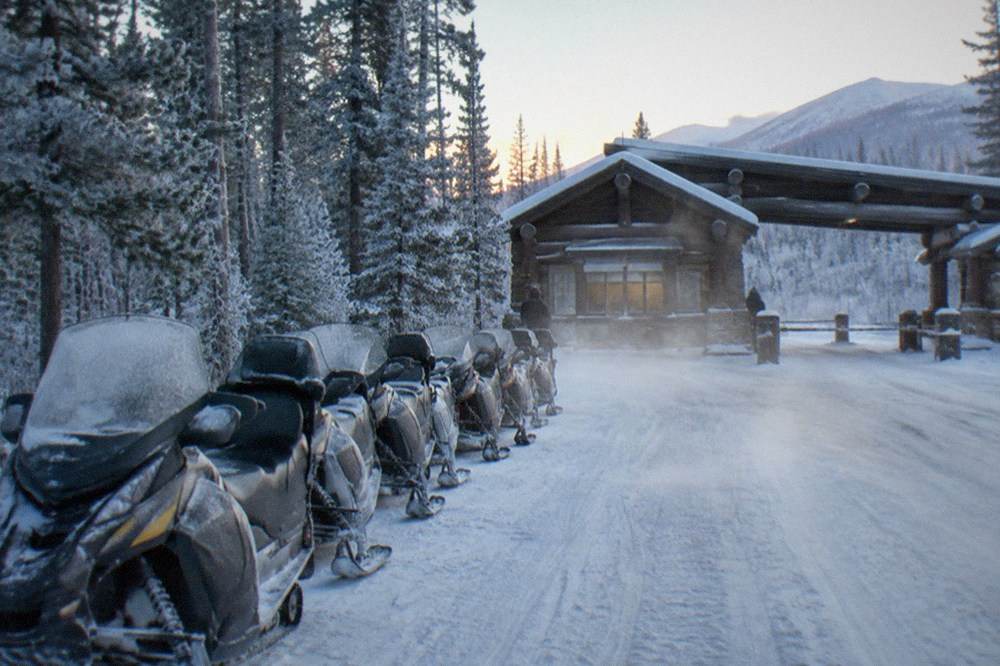 A line of parked snowmobiles at a snowy forest trailhead at dawn