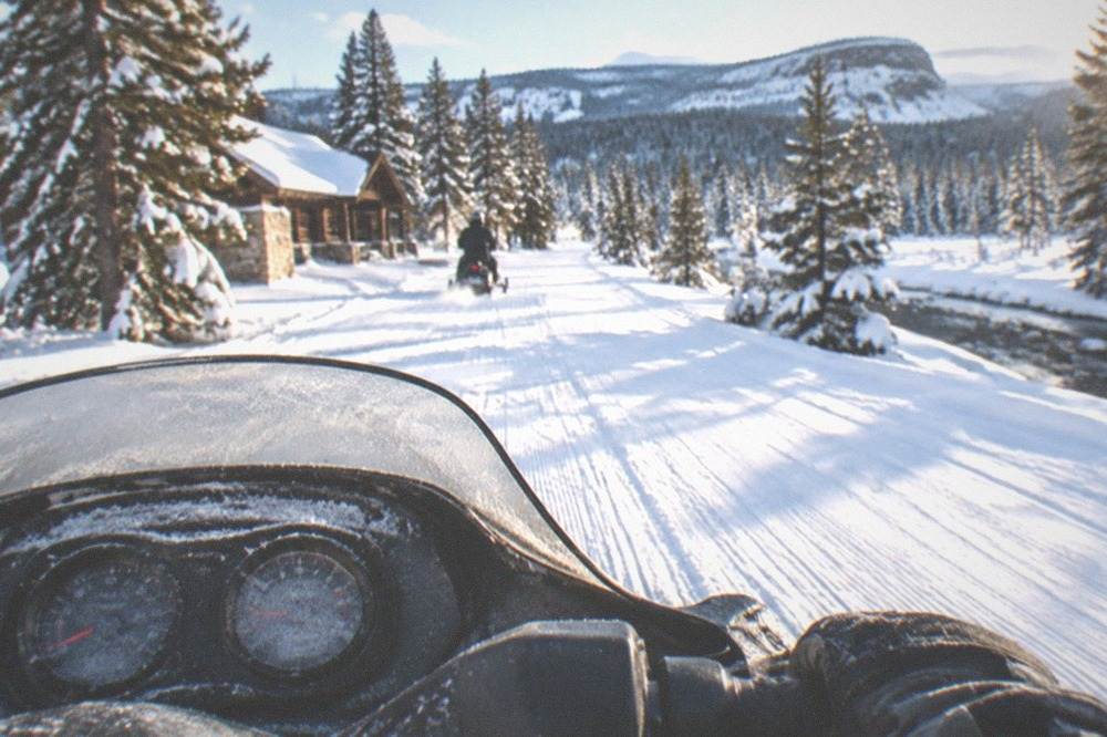 A snowmobile dashboard and gloved hands on a wide white groomed road through pine forest