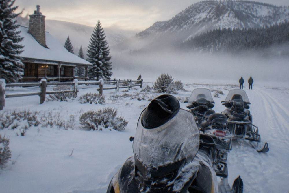 Snowmobiles parked on a snowy meadow edge below forested mountains