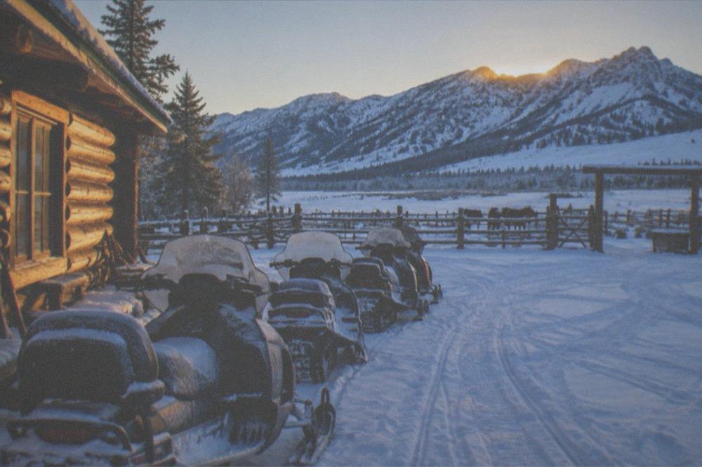 A line of parked snowmobiles on a snowy ranch meadow beside a log lodge at dawn