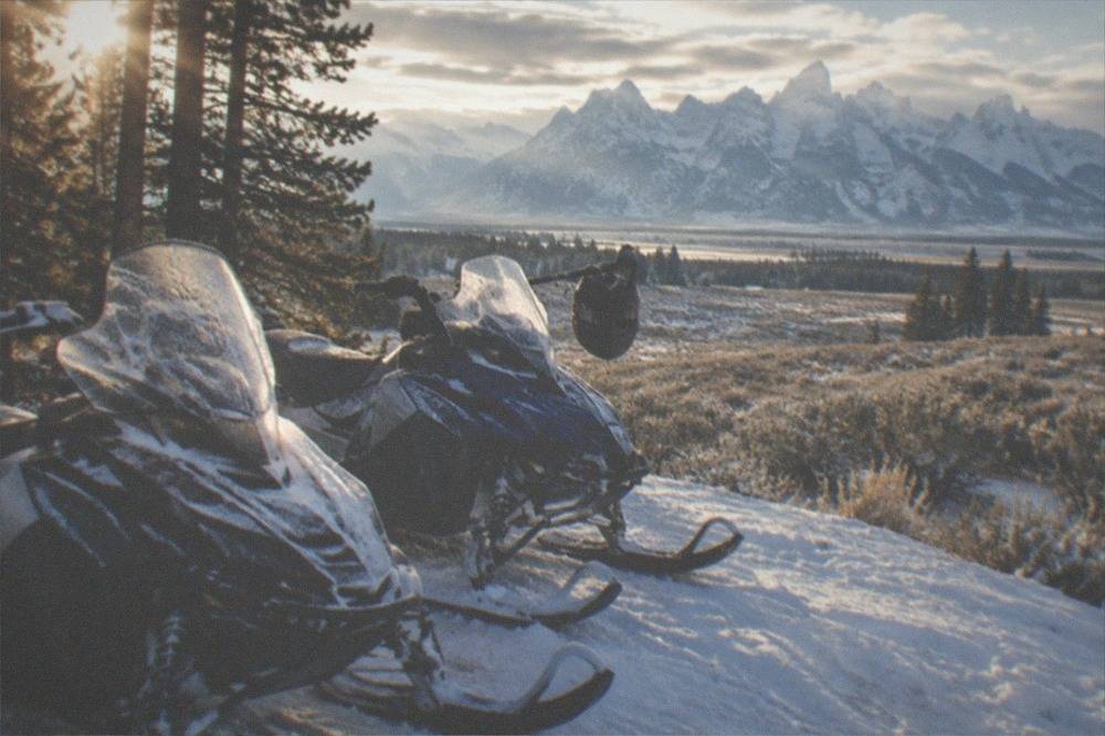 Two snowmobiles parked on a snowy forest trail with tall peaks behind them