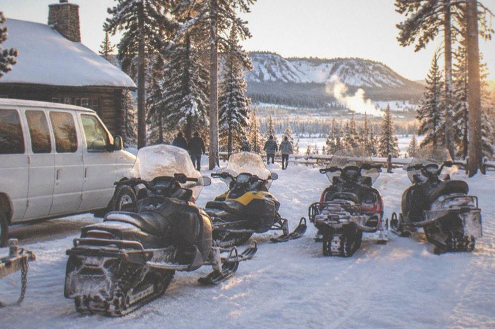 Van and snowmobiles staged at a winter trailhead beside snow-laden trees at sunrise
