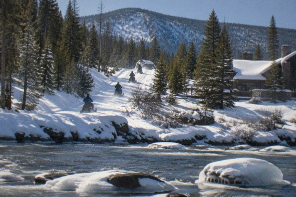 A line of snowmobiles traveling along a snow-covered park road past steaming thermal ground