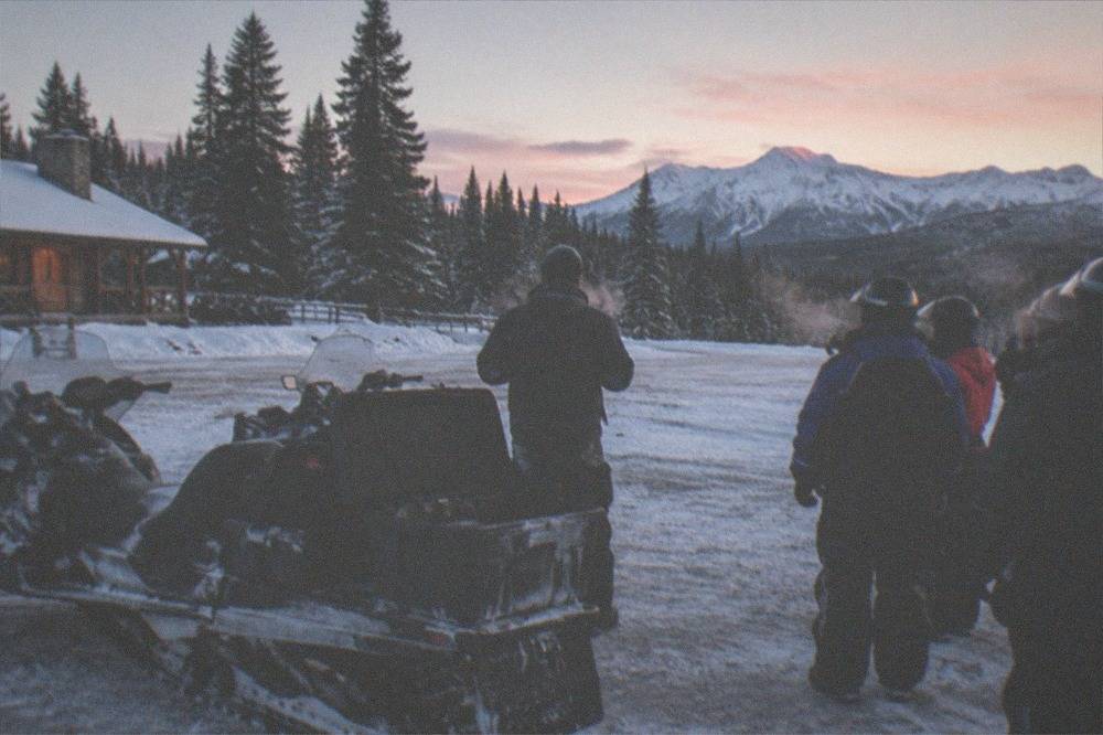 A group of riders in snowmobile suits listening to a guide beside parked sleds at a snowy trailhead