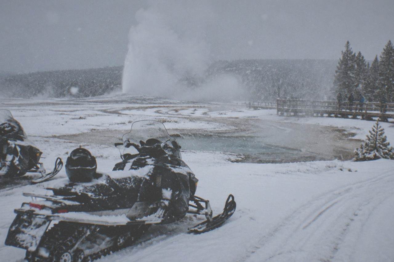 Snowmobiles parked on a groomed white road beside steaming geothermal pools in winter