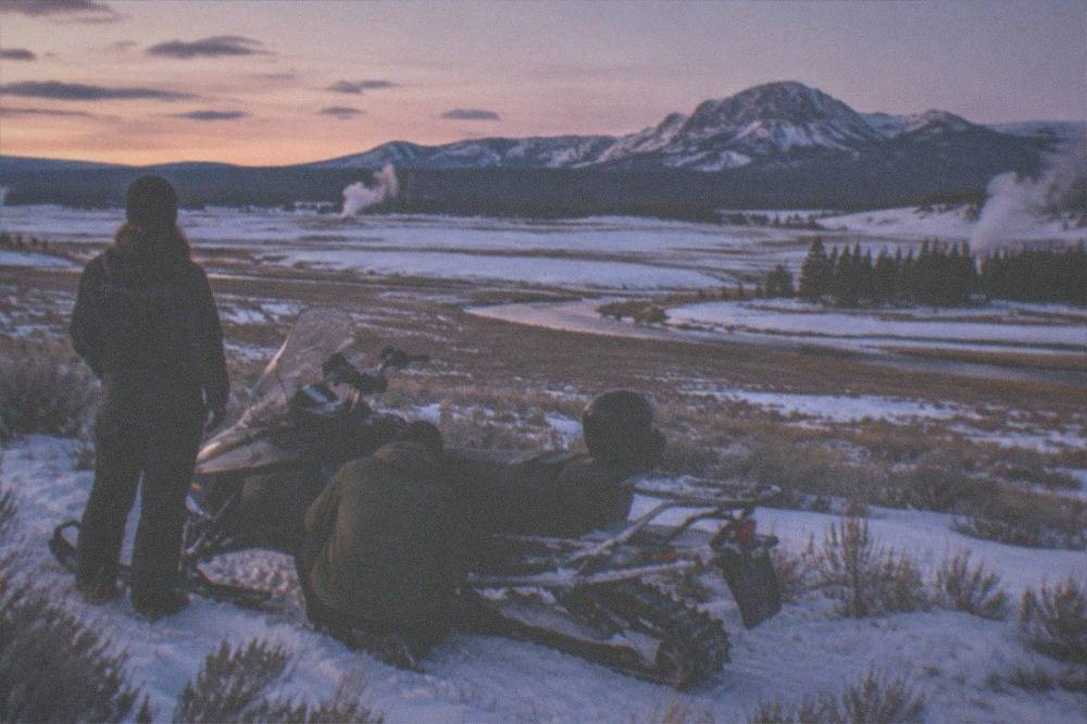 A snowmobile guide coaching a rider beside a parked sled in an open snowy meadow