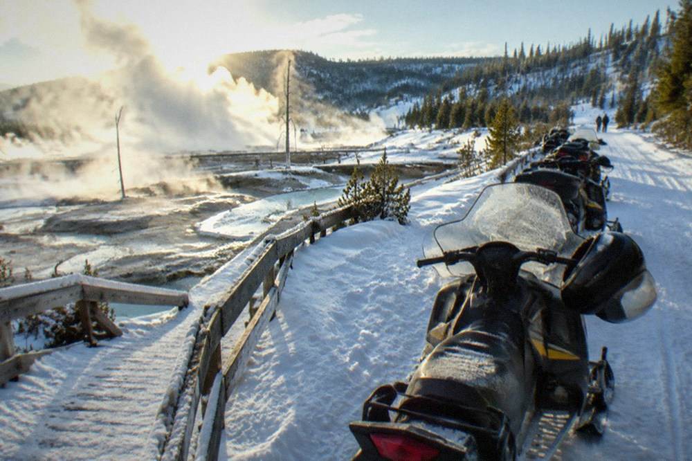 A line of snowmobiles parked on a snowy road beside steaming geothermal pools in a wide geyser basin