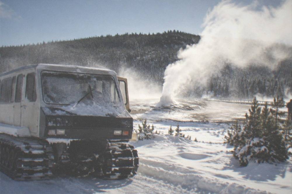 A tracked snowcoach vehicle parked on a snow-packed road beside steaming fumaroles