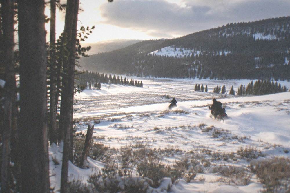 Two snowmobiles climbing a wide open snowy meadow beneath forested hills in afternoon light