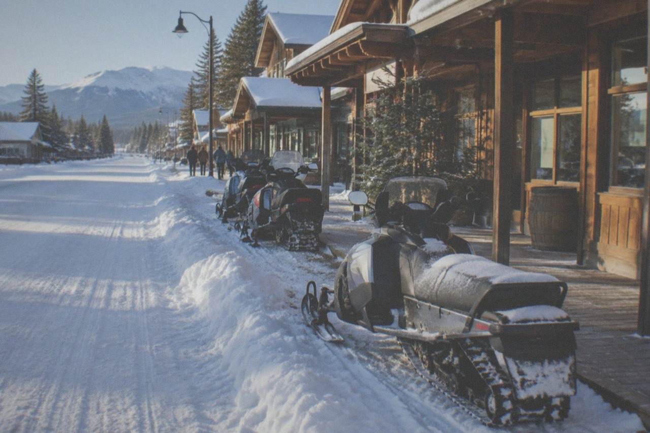 Snow-covered street in a small gateway town with snowmobiles parked along the storefronts under a clear winter sky