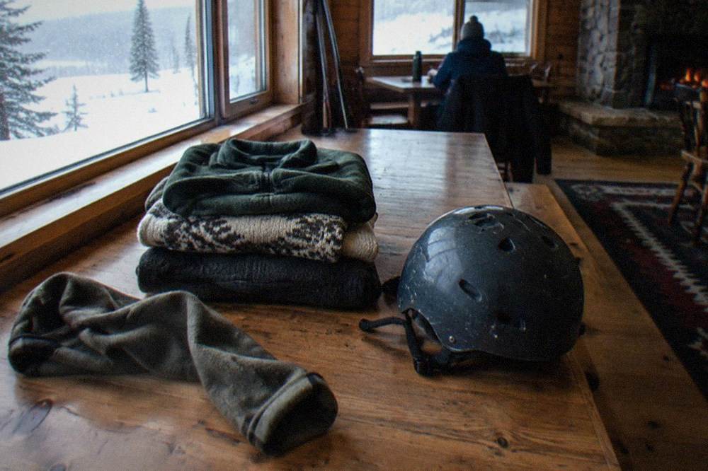 Fleece jackets and wool base layers folded in a stack beside a snowmobile helmet on a wooden table