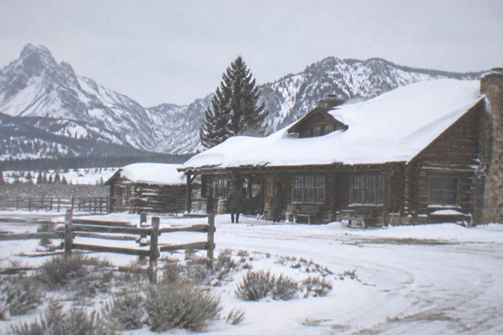 A wooden ranch lodge with deep snow on the roof and a valley behind it