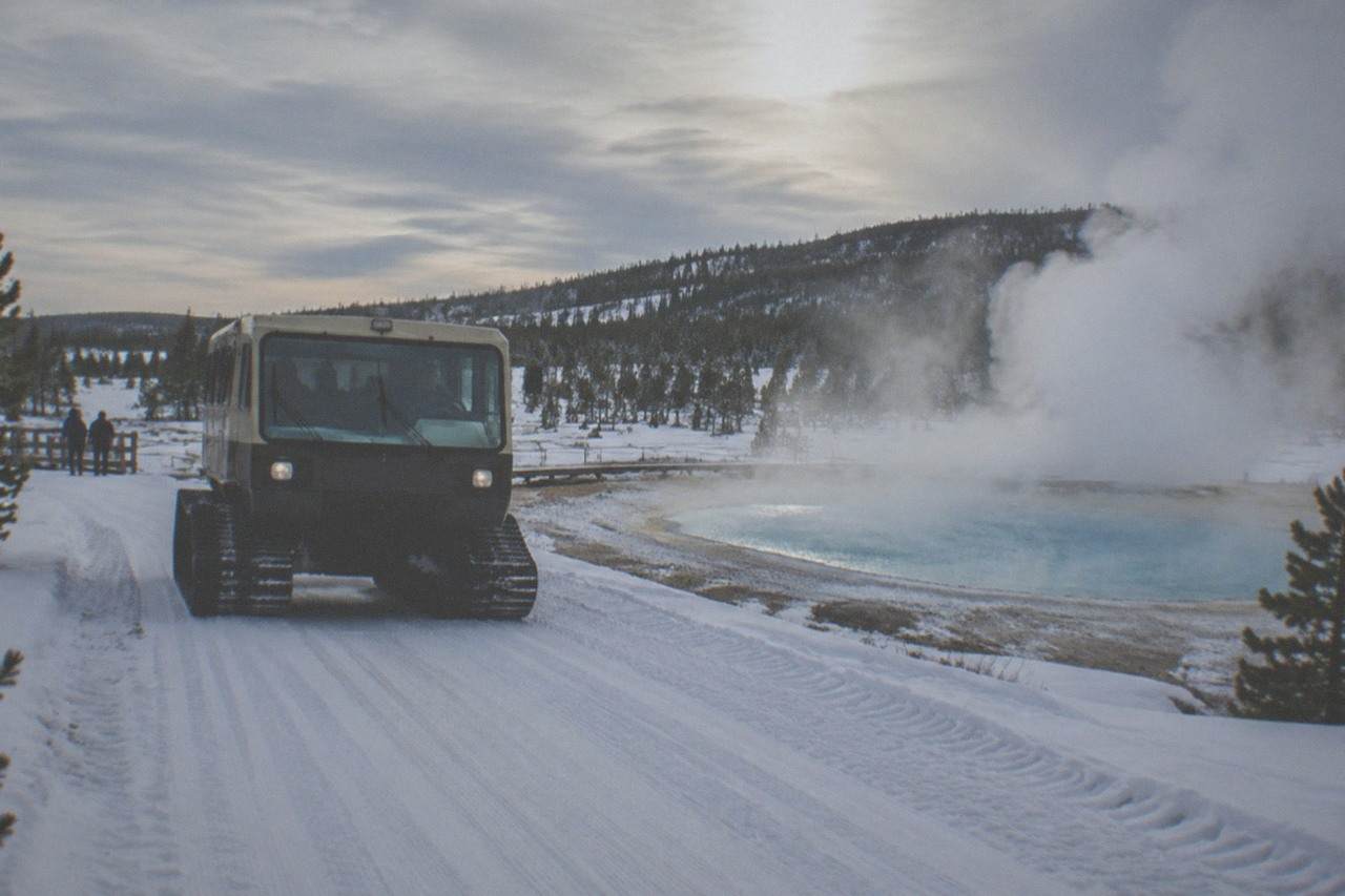 A heated snowcoach traveling a groomed snowy road past steaming thermal features in Yellowstone