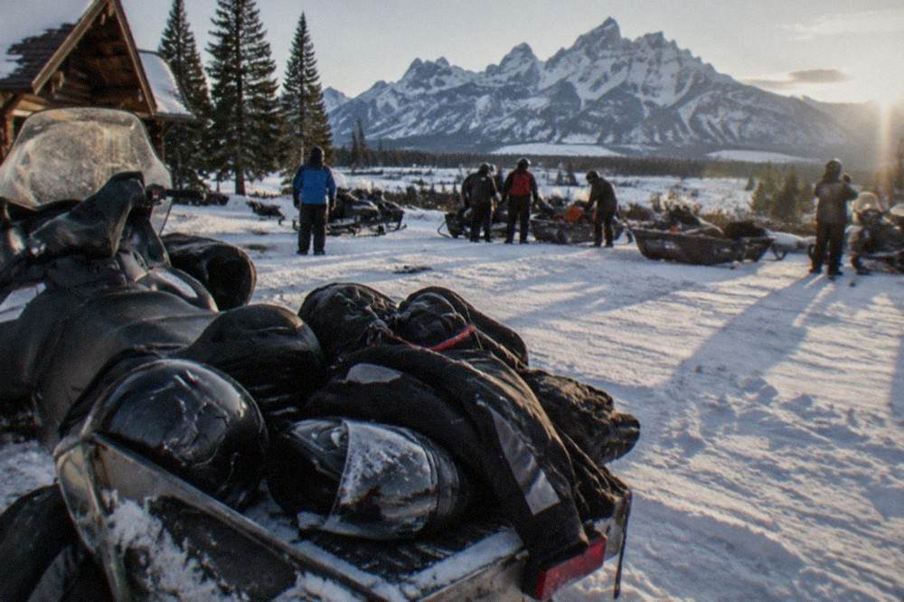 Snowmobile helmet and insulated gear laid out beside a parked snowmobile on a snowy trailhead