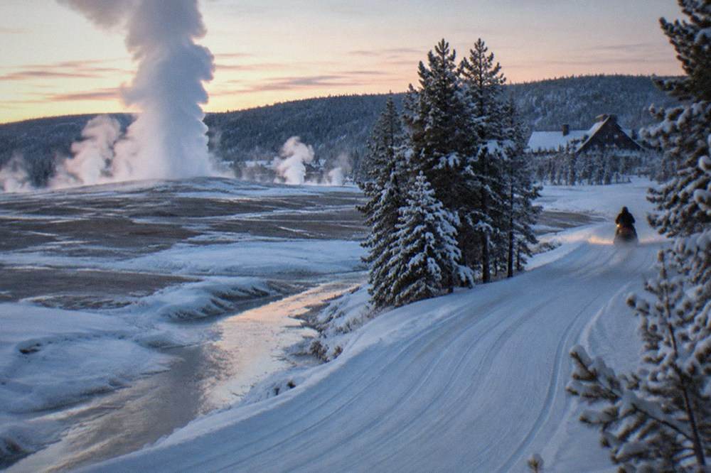Steam rising from a snowy geyser basin with a groomed winter road curving alongside