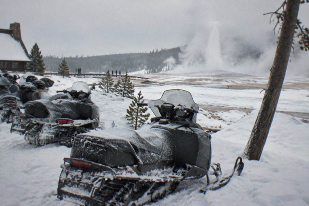 Snowmobiles parked in a line beside a steaming geyser basin under a pale winter sky