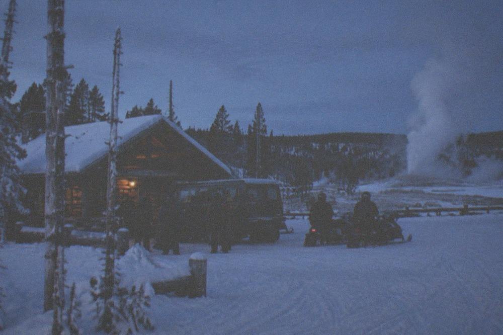 Snowcoach and snowmobile groups gathered at a warming hut with steam rising in winter dusk light